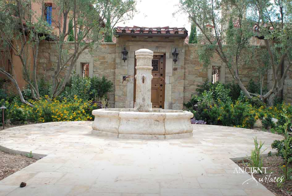 Round reclaimed limestone pool fountain with a tall square pillar and iron spouts standing at the centre of a paved walled courtyard
